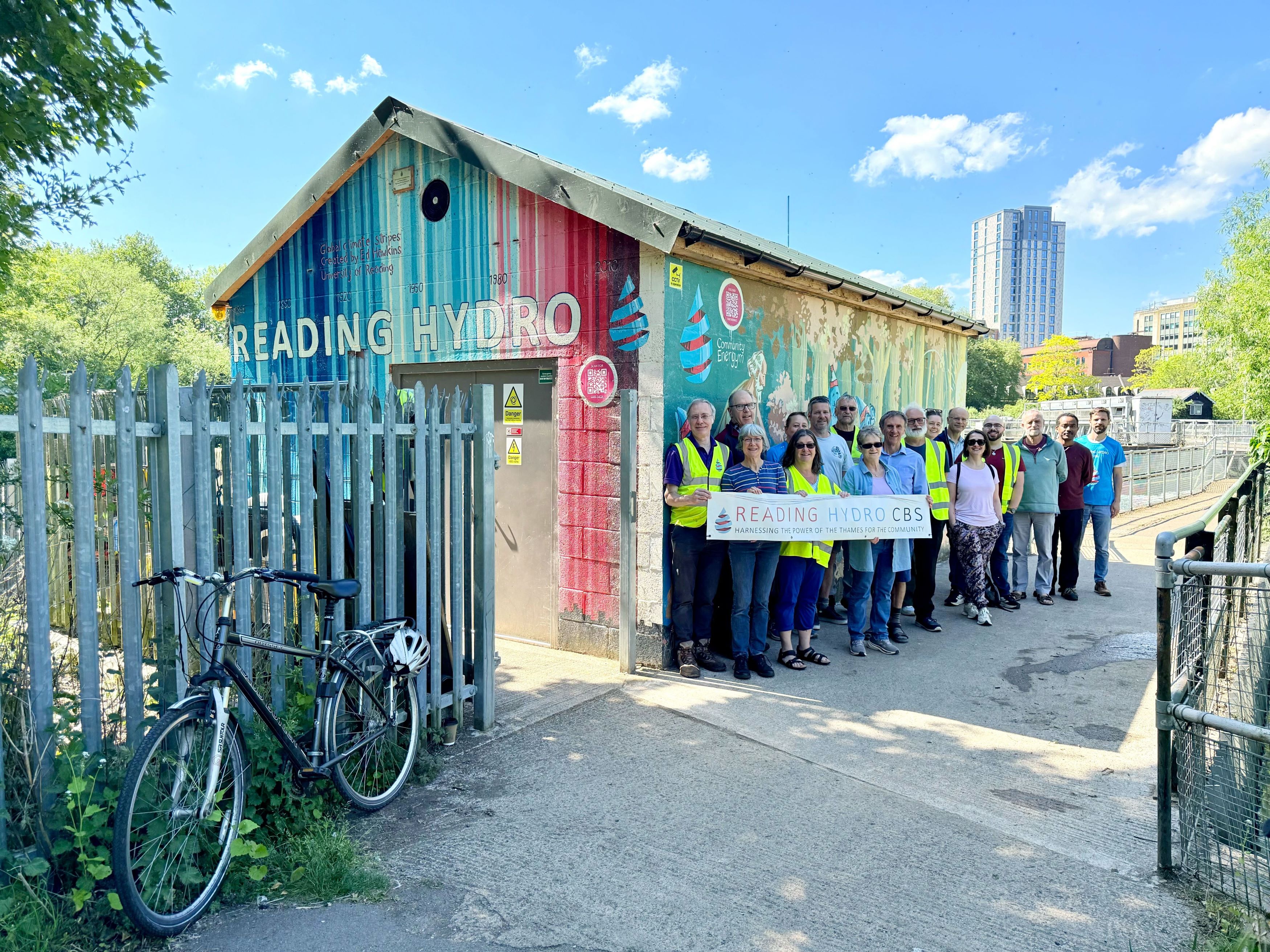 A group of people standing in front of the Reading Hydro turbine house, holding a sign that reads 'Reading Hydro CBS'. There is a bicycle parked to the left of the group.